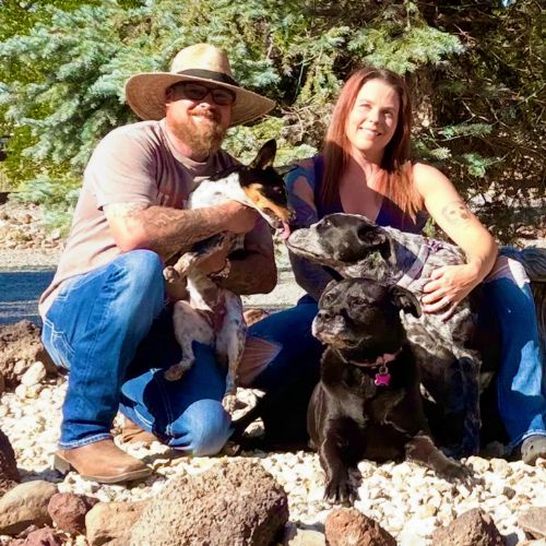 Two people crouch outdoors with a group of dogs on a gravel surface,